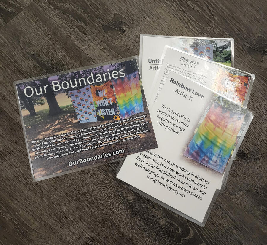 A pile of laminated paper signs, three of which show artist's statements for individual works, and one of which is an overall explanation of the project. a photo of all three current barriers is the background